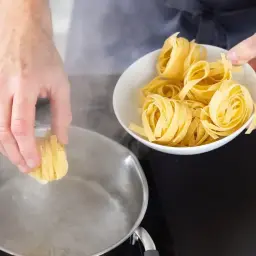 Ręka wkłada porcję makaronu do gotującej się wody. W misce obok czeka kolejna porcja, by uzyskać idealny makaron al dente.
