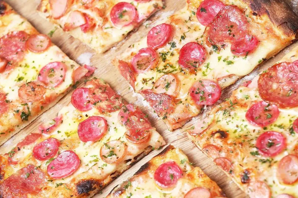 Kawałki pysznej pizzy na cienkim cieście z kiełbasą i serem, posypane ziołami.